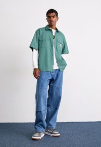 Camisa verde de manga curta com dois bolsos no peito, sobre uma camisa branca de manga longa. Calças de ganga azuis largas e ténis cinzas.