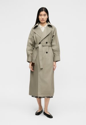 Kobieta ubrana w długi beżowy trencz z paskiem, czarne baleriny i koronkową jasną bluzkę pod płaszczem, stojąca na jednolitym tle.