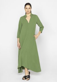 Zielona długa sukienka z dekoltem w serek, rękawami o d�ługości trzy czwarte, asymetrycznym dołem i boczną kieszenią, uszyta z lekkiego materiału. Noszona z czarnymi sandałami.
