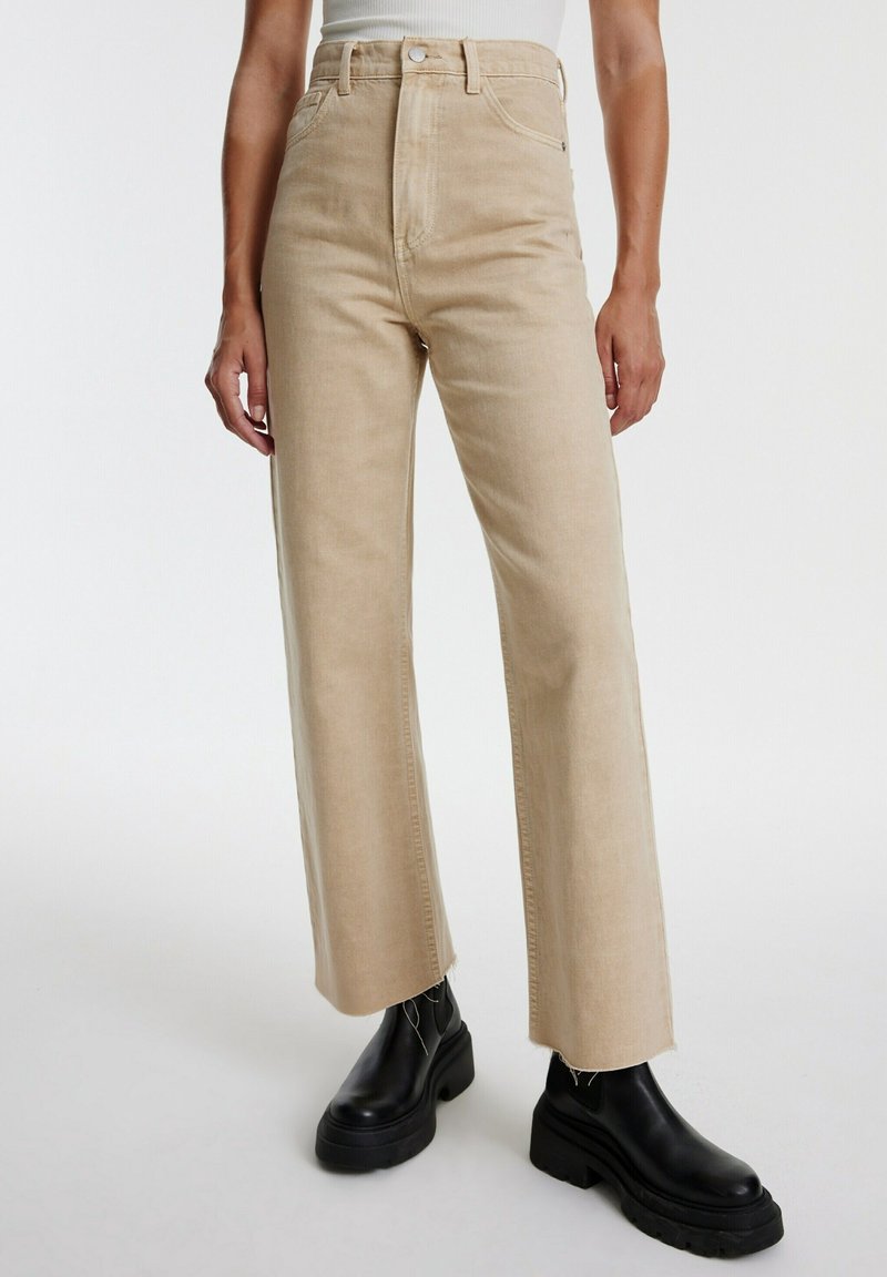 Pantalon beige taille haute à jambes droites avec ourlets bruts, porté avec des bottines noires épaisses à plateforme et un top blanc sans manches.