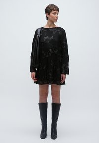 Μαύρο φόρεμα με κεντήματα λουλουδιών, μακριά μανίκια και φουσκωτή σιλουέτα. Συνδυάζεται με μαύρες μπότες μέχρι το γόνατο και μια τσάντα ώμου με υφή.