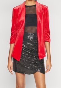 Femme portant une veste en velours rouge, un haut noir transparent et une mini-jupe noire à sequins, vue des épaules jusqu'à mi-cuisse.
