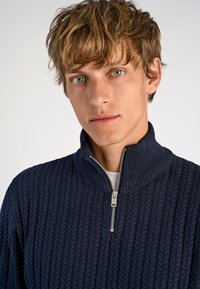 Navyblaue Strickjacke mit hohem Kragen und halber Reißverschluss; hat ein strukturiertes Muster und gerippte Bündchen.