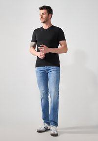 Tricou negru cu mânecă scurtă și decolteu în V, blugi albaștri și adidași albi cu accente negre, purtate de un bărbat care stă în picioare cu mâinile încrucișate.