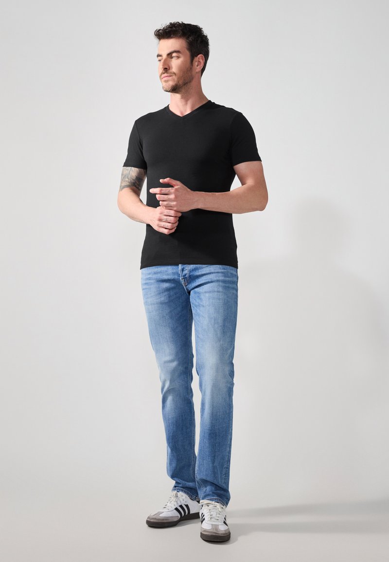 Tricou negru cu mânecă scurtă și decolteu în V, blugi albaștri și adidași albi cu accente negre, purtate de un bărbat care stă în picioare cu mâinile încrucișate.