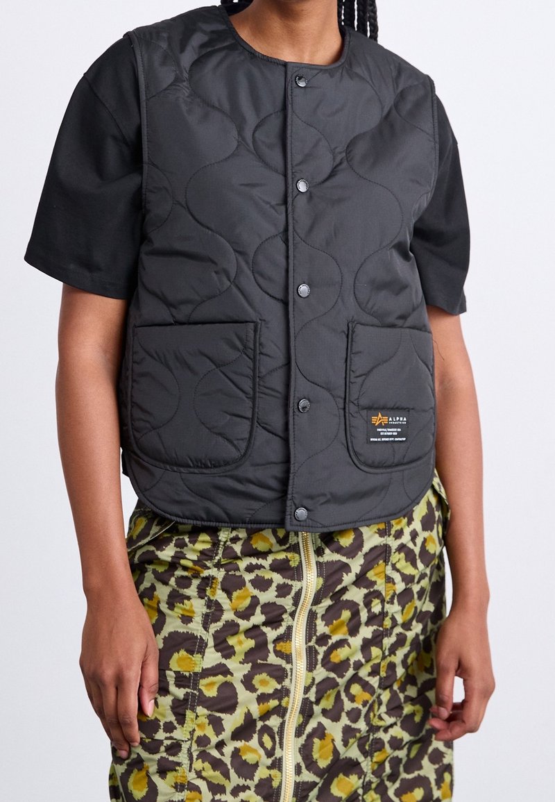 Personne portant un gilet matelassé noir avec boutons-pression par-dessus un t-shirt noir à manches courtes et une jupe verte à imprimé léopard avec fermeture éclair devant.
