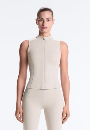 Femme portant un haut zippé sans manches beige et un pantalon assorti ajusté, debout devant un fond blanc uni.