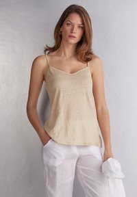 Beige camisole met dunne bandjes, v-halsontwerp, gestructureerde stof. Gecombineerd met witte linnen broek, model houdt een witte accessoire vast.