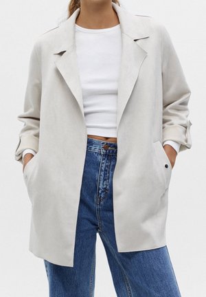 Vrouw draagt een lichtbeige lange blazer over een wit cropped topje en een blauwe high-waist jeans, handen in de zakken, neutrale achtergrond.