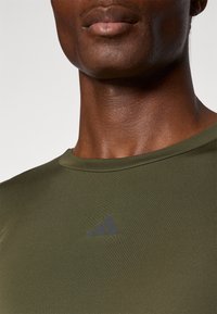 Olivově zelené sportovní tričko s kulatým výstřihem, s jemným černým logem Adidas na hrudi, vyrobené z hladkého, přiléhavého materiálu.