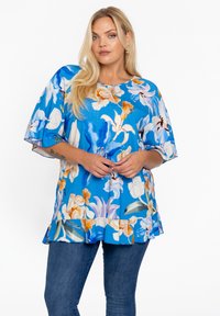 Bloemenblouse in helderblauw met korte mouwen, een ronde halslijn en een rucheszoom, versierd met gele en witte bloemenpatronen.