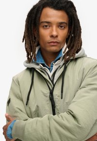 Lichtgroene bomberjack met zwarte hardware, grijze hoodie eronder en een blauwe denim shirt. Heeft een relaxte pasvorm en een trekkoord in de capuchon.