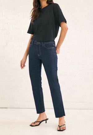 Femme portant un t-shirt noir à manches courtes, un jean droit taille haute bleu foncé et des sandales noires à talons ouvertes, debout à l'intérieur.