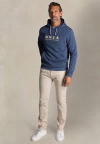 Blauwe katoenen hoodie met witte print "N.Z.A. New Zealand Auckland" en trekkoord. Gecombineerd met lichtbeige broek en witte sneakers.