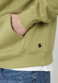Sweatshirt vert olive avec une poche avant, des poignets côtelés et un petit logo noir. Porté sur un T-shirt blanc et un jeans en denim bleu.