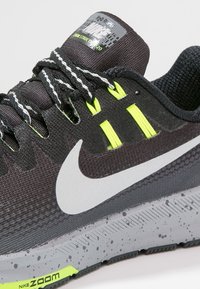 Chaussure de sport noire avec des accents gris et vert lime. Comprend une tige en maille respirante, un logo réfléchissant et une semelle texturée avec des éclats.