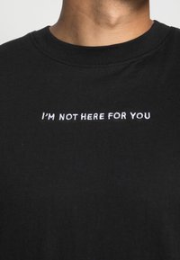 Svart bomullströja med vitt broderat texten "JAG ÄR INTE HÄR FÖR DIG" på framsidan. Rund halsdesign med korta ärmar.