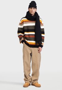Pull en maille rayé dans des tons terreux variés, écharpe noire, pantalon en velours côtelé beige et chaussures marron clair. Le modèle se tient devant un fond neutre.