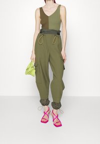 Body in Jersey verde oliva con costine e scollatura a V, abbinato a pantaloni joggers verdi oliva di vestibilità ampia e tacchi rosa con cinturini. Porta una piccola borsa verde.