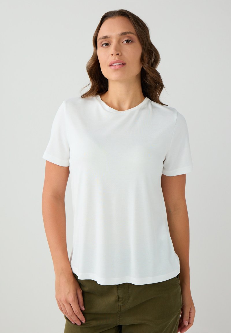 Vrouw met golvend bruin haar, gekleed in een effen wit T-shirt met korte mouwen en olijfgroene broek, staande tegen een lichtgrijze achtergrond.