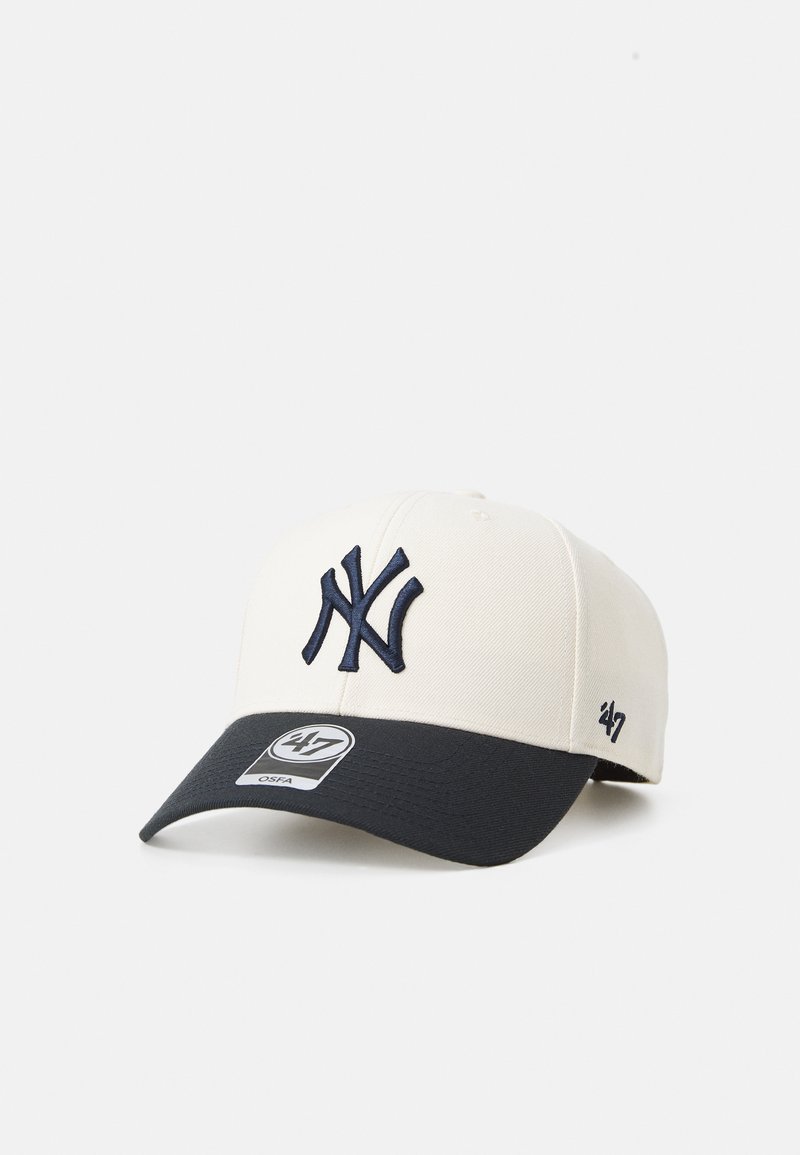 Krämfärgad basebollkeps med en marinblå skärm, som har en broderad NY-logotyp och en tagg som visar märket "47" längst fram.
