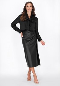Fekete selyem hosszú ujjú blúz, masnival, fekete műbőr midi szoknyával párosítva, oldalsó hasítékkal és sima felülettel.