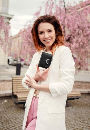 Černá peněženka s kovovým akcentem, obdélníkový tvar, s vyraženým nápisem "VUCH," kterou drží žena v bílém saku a růžových kalhotách.