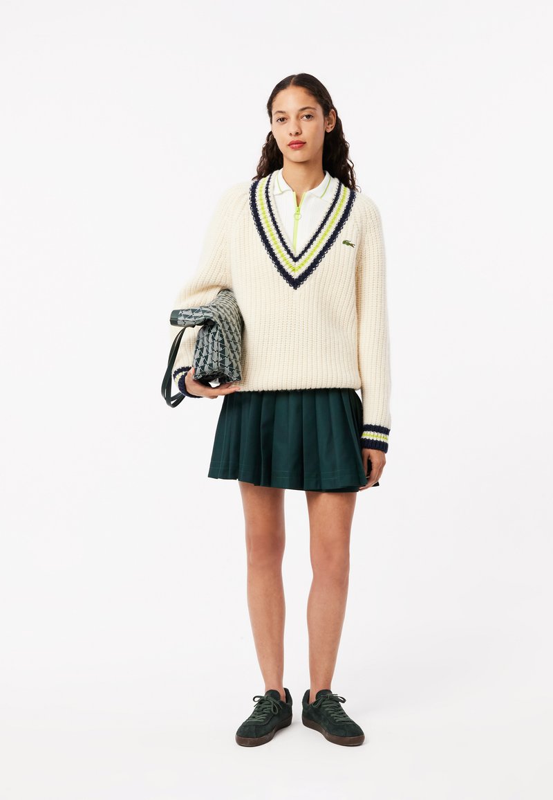 Ivoryfarbener, gestrickter Pullover mit V-Ausschnitt und blau-gelben Streifen, kombiniert mit einem dunkelgrünen, gefalteten Rock und grünen Sneakers, während sie eine gemusterte Tasche hält.