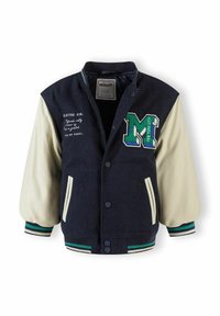 Tumesinine ülikooli jakk kreemjate varrukatega, millel on roheline broderi „M“ patch ja triibulised soonikud. Ees nupud ja taskud.