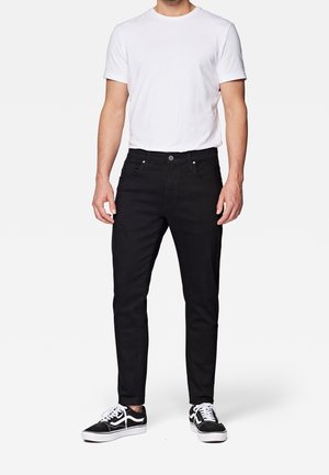 Hombre con camiseta blanca de manga corta lisa, vaqueros ajustados negros y zapatillas negras y blancas, de pie frente a un fondo liso.