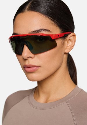 Donna con capelli scuri che indossa occhiali sportivi avvolgenti con montatura rossa e un top sportivo color taupe, leggermente rivolta a sinistra su uno sfondo bianco.