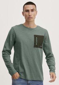 Langarmshirt aus weichem grünem Stoff, mit einer dunkelgrünen Brusttasche mit Reißverschluss und einem kleinen Logotag.