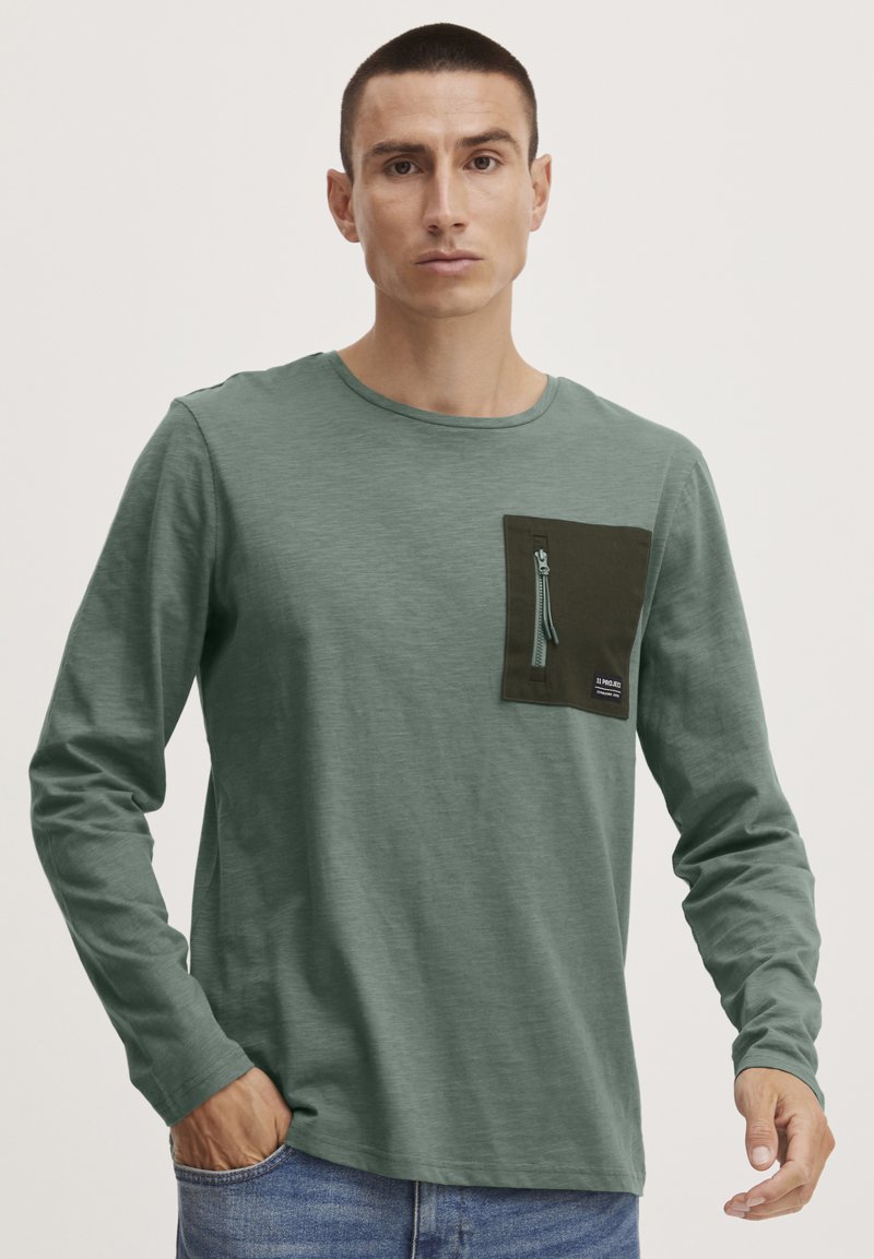 Langarmshirt aus weichem grünem Stoff, mit einer dunkelgrünen Brusttasche mit Reißverschluss und einem kleinen Logotag.