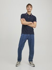 Mørkeblå polo shirt med rød og hvid stripekrave, parret med lyseblå jeans og grå sneakers, stående med armene krydset mod en ensfarvet baggrund.