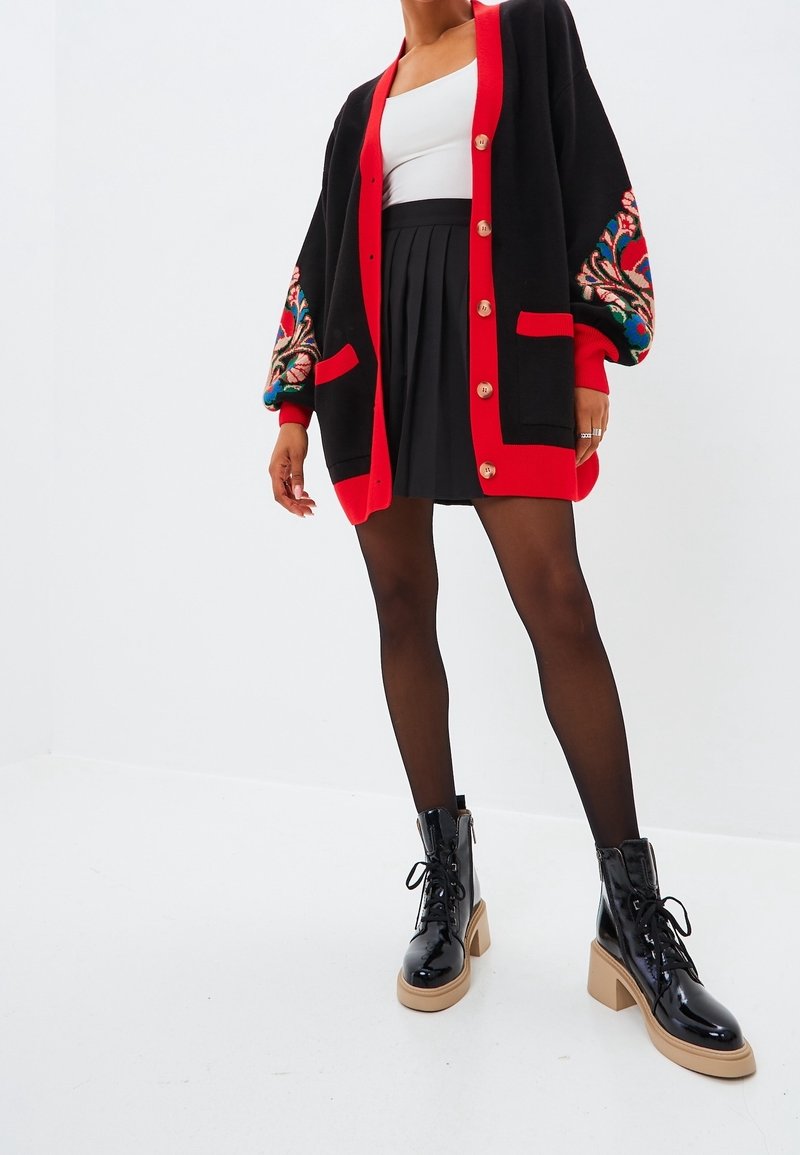 Black cardigan with red trim and floral embroidery, paired with a black pleated skirt and black patent leather ankle boots.