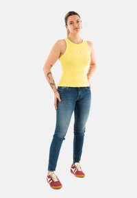 Top de tirantes amarillo con estrías, vaqueros ajustados azules y zapatillas retro burdeos con acentos blancos. Un atuendo casual con un tatuaje visible en el brazo.