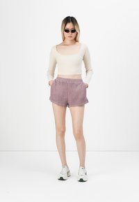 Crop-top beige à manches longues, shorts lilas texturés avec poches latérales, et baskets blanches avec accents verts. Mannequin portant des lunettes de soleil noires.