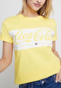 Żółta bawełniana koszulka z białym poziomym paskiem z logo "Coca-Cola" oraz "TOMMY JEANS". Krótkie rękawy, luźny krój, prosty design.