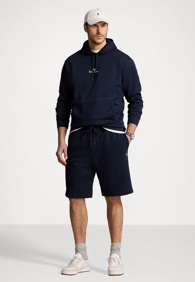 Tamsiai mėlynas džemperis su priekinėmis kišenėmis ir raišteliu, atitinkančios tamsiai mėlynos spalvos šortai, balta kepurė ir balti sportbačiai su smėliniais akcentais. Tekstūruota medžiaga.