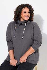 Femme souriante aux cheveux bouclés portant un pull gris avec des cordons blancs, assise et tenant l'ourlet du pull des deux mains.
