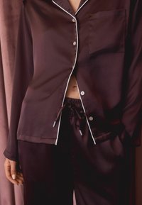 Ciemnobrązowy satynowy komplecik piżamowy, składający się z koszuli z guzikami z białymi lamówkami i kieszonką na piersi, w komplecie z dopasowanymi spodniami i ściągaczem w pasie.