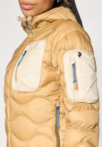 Beige pufferjack met een gestructureerd, gewatteerd ontwerp, contrasterende crèmekleurige zakken, blauwe accenten en functionele ritsen.