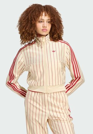 Junge Frau mit lockigem Haar, die eine cremefarbene und rot vertikal gestreifte Trainingsjacke und passende Hose trägt, steht mit den Händen hinter dem Rücken.