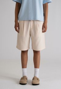 Beige pleated shorts with side pockets, paired with a light blue t-shirt and beige slip-on shoes, highlighting a relaxed fit and casual style.