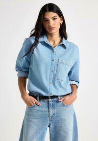 Lyseblå denimskjorte med knaplukket krave, brystlomme og opkørte ærmer, parret med højtaljede lyseblå jeans.