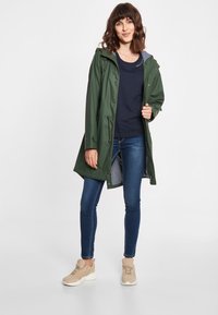 Groene waterdichte jas met een capuchon en ritssluiting, met een gestreepte voering aan de binnenkant. Gecombineerd met een marineblauw shirt en blauwe jeans, vergezeld van beige sneakers.