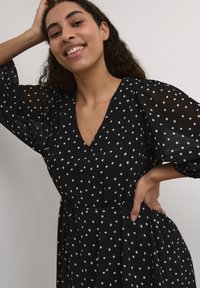 Vestido negro con lunares hecho de una tela ligera, que presenta un escote en V profundo, mangas abullonadas transparentes y un detalle de cintura atada.