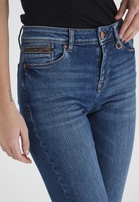 Jeans ajustados azul oscuro de mezclilla, con un bolsillo con cremallera para monedas, detalles en metal plateado y un sutil efecto de desgastado en la parte frontal.