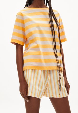 Kobieta ubrana w luźną, żółto-białą koszulę w paski z krótkim rękawem i dopasowane spodenki w paski, z długimi splecionymi warkoczami spadającymi na ramię.