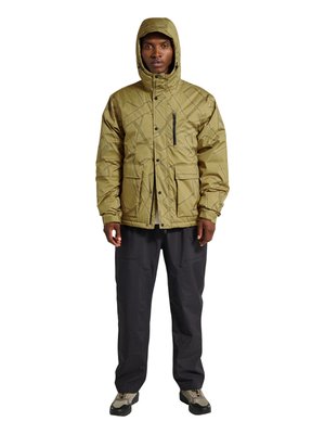 Homme portant une veste matelassée à capuche vert olive, un pantalon noir et des chaussures de randonnée beiges, debout devant un fond blanc.